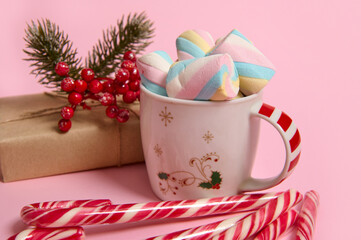 Close-up of coffee mug with colorful marshmallows, Christmas gift in craft wrapping paper, holly and striped candy canes on pink pastel background. Christmas, New year concept with copy space for ad