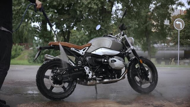 Young man washing his scrambler motorbike on the street. Cleaning cafe racer with a water hose. A guy waters motorcycle. High quality FullHD footage