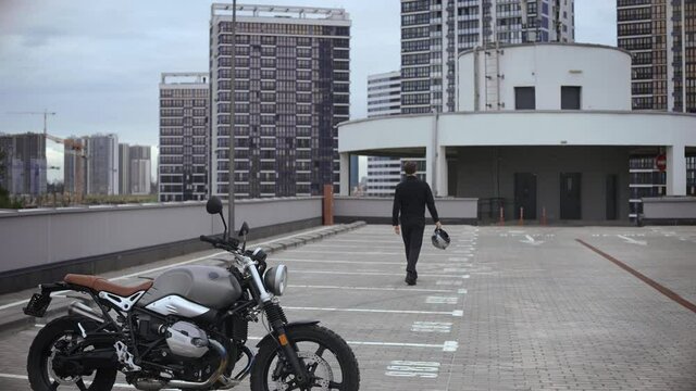 Young man is coming away from scrambler motorbike on top of the multi-level parking in city. Guy leaving cafe racer in empty parking garage. High quality 4k footage