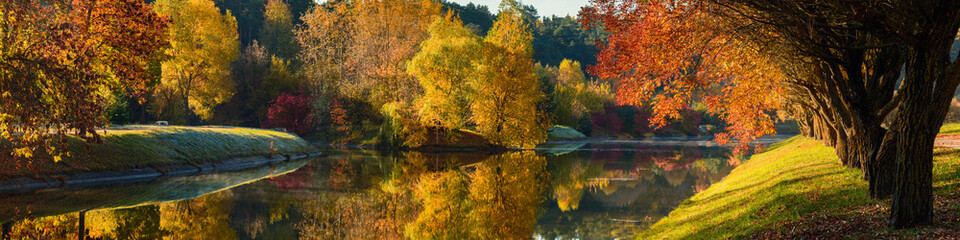 vibrant colors of October. wide panoramic view of the yellow-orange autumn morning park with lush trees reflected in the river water. picturesque fall landscape