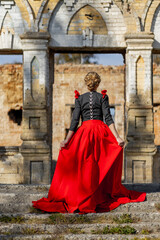 Fototapeta na wymiar A woman in a 19th century dress in red and black walks to the ruins of an old castle manor in autumn
