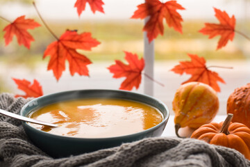 A bowl of pumpkin soup with mini pumpkins by a window with maple leaves