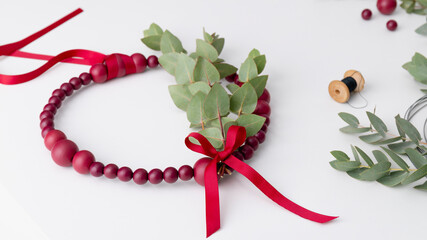 Decorative wreath made of red beads and eucalyptus on the white table. Workspace with beads and threads.