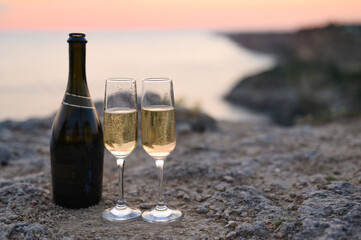 Romantic evening on the background of the sunset. Two glasses of champagne with an open bottle on the edge of a cliff against the backdrop of the sea and sunset. Selective focus on glasses