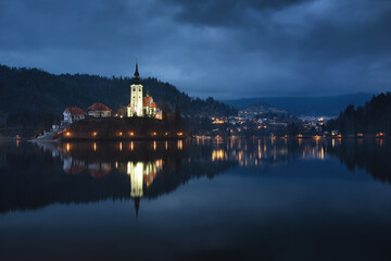 Fototapeta na wymiar A mysterious evening view of Lake Bled, Slovenia. Landscape with the lights of the city at night. Church on an island, postcard view, national park.