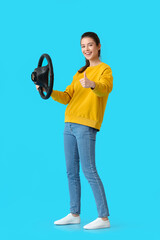 Young woman with steering wheel showing thumb-up on color background