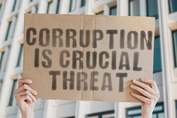 The phrase " Corruption is crucial threat " on a banner in men's hand with blurred background. Politics. Government. Dangerous. Economy. Finance. Warning. Dishonesty. Trick. Corrupted