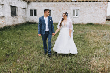 Stylish groom in a blue suit and a brunette bride in a white lace dress stroll through the village in nature on the background of a brick building, a farm. Wedding photography, portrait.