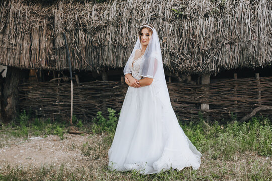 A beautiful, sweet bride in a white dress with a veil and a diadem on her head, standing in the village against the background of a hut with a straw. Wedding photography, portrait.