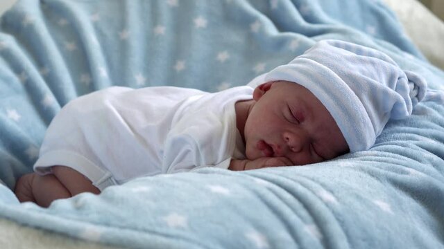 Close-up Newborn Baby Face Portrait Early Days Sleeping Sweetly On Tummy Blue Star Background. Child At Start Minutes Of Life on Hat. Infants, Childbirth, First Moments Of Borning, Beginning Concept