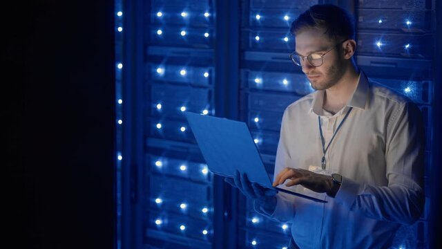 Data center IT specialists coworking in secure server room of cyberspace. Male administrator pointing at error server cabinet running diagnostics on laptop computer