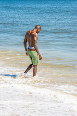 A well built young sexy black guy, half naked, whole body wet,  is walking into water of ocean, huge rolling splash waves. .