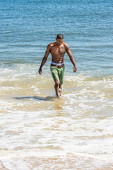 Fototapeta na wymiar A well built young sexy black guy, half naked, is walking on water of ocean, looking down, smiling. .