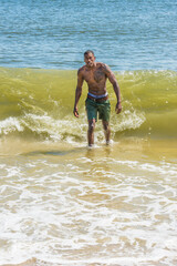 A well built young sexy black guy, half naked,  is confidently walking out off ocean. Huge waves coming from back..