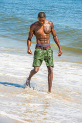 A well built young sexy black guy, half naked and whole boy wet,  is looking down and walking on the beach..