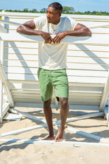 Fototapeta premium Bending over, both arms resting on a stick, a young handsome black guy is standing by a white wooden structure on the beach, looking around, relaxing..
