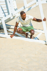 Wearing a white Henley shirt, green shorts, barefoot, a young handsome black guy is squatting under a white wooden structure on the beach, bending over,  looking around.