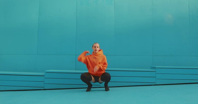 Dancing homosexual man wearing heels performing vogue femme dance outdoors by blue wall. Gender and art concept