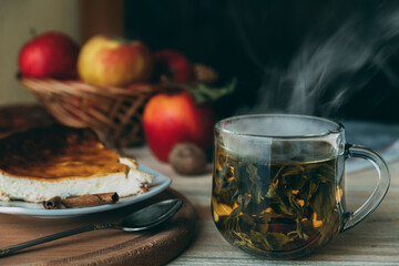 Glass cup with green tea from which steam comes out on a wooden table. Next to it is a cheese casserole on a white plate. In the background is a basket with apples and nuts.