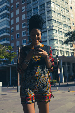Beautiful African American woman wearing casula red and black dress and african turban standing up texting on the mobile cell phone and smiling. Palm trees and city buildings on the background.