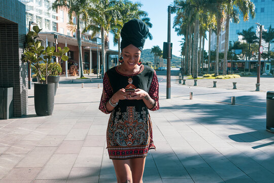 Beautiful African American woman wearing casula red and black dress and african turban standing up texting on the mobile cell phone and smiling. Palm trees and city buildings on the background.