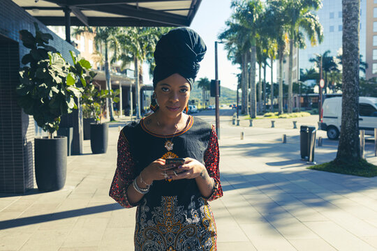 Beautiful African American woman wearing an african turban standing up texting on the mobile cell phone and looking at the camera. Palm trees and city buildings on the background.