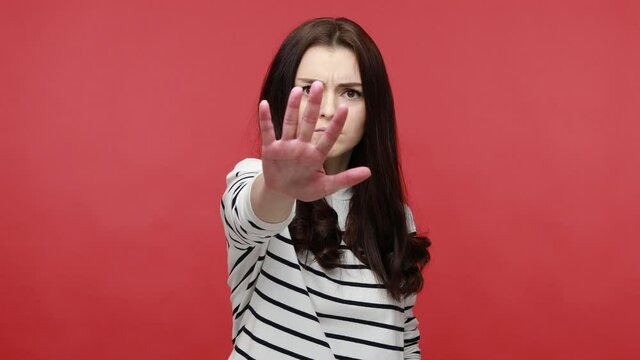 Young woman making stop gesture with her palm outward, saying no, expressing denial or restriction, wearing casual style long sleeve shirt. Indoor studio shot isolated on red background.