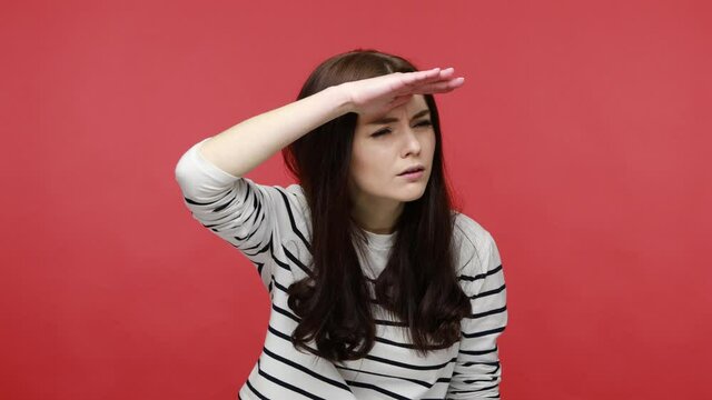Woman keeps palm over head, looking attentively far away, peering with expectation at long distance, wearing casual style long sleeve shirt. Indoor studio shot isolated on red background.