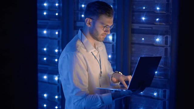 Data center IT specialists coworking in secure server room of cyberspace. Male administrator pointing at error server cabinet running diagnostics on laptop computer