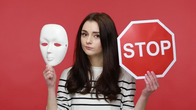 Portrait of attractive woman holding white mask and stop traffic sign to camera, nodding her head no, wearing casual style long sleeve shirt. Indoor studio shot isolated on red background.