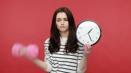 Woman holding wall clock and pink dumbbell, it is time for sport, calls on to active lifestyle, wearing casual style long sleeve shirt. Indoor studio shot isolated on red background.