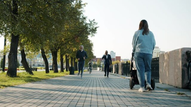 Diverse group of pedestrians are moving along urban street enjoying summer city doing sports walking with baby carriage and riding bike