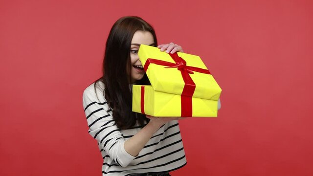 Portrait of curious woman opens gift box, looking inside, being disappointed to recive sush present, wearing casual style long sleeve shirt. Indoor studio shot isolated on red background.