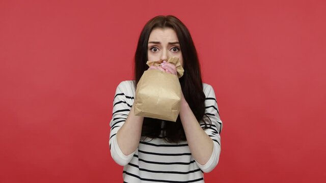Portrait of woman hardly breathing using paper bag, trying to calm down, to improve well-being, wearing casual style long sleeve shirt. Indoor studio shot isolated on red background.