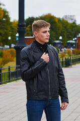 Young man in a black leather jacket and jeans in a park area. Opens the zipper. Yellowed plants in blur in the background.