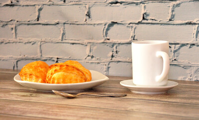Curd buns on a white plate and a cup of black coffee on a wooden table against a brick wall.