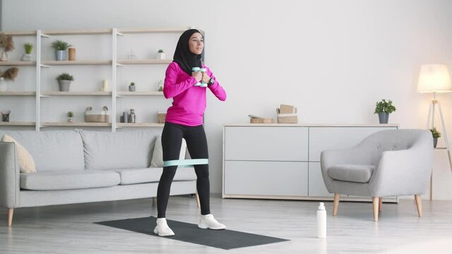 Young active muslim lady wearing hijab making deep squats exercise, using elastic band and dumbbells at home