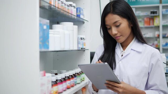 Asian pharmacists working and checking drug in pharmacy store at hospital. Asian doctor checking medicine cabinet and wearing white medical coat. Shelves pharmacy background in drugstore.