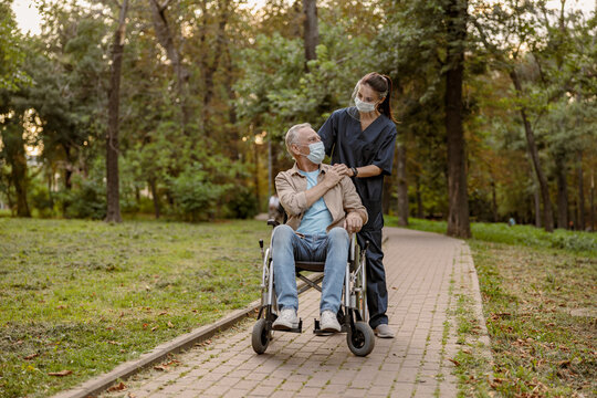 Full Length Shot Of Young Nurse In Face Shield And Protective Face Mask Supporting Mature Handicapped Man In Wheelchair During A Walk In The Park