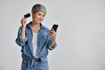 Fototapeta premium Portrait of middle aged Asian woman 50s wearing casual denim shirt white t-shirt holding smartphone and credit card shopping online isolated on white background