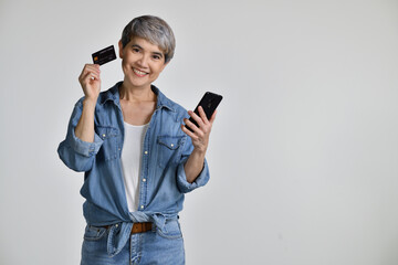Portrait of middle aged Asian woman 50s wearing casual denim shirt white t-shirt holding smartphone and credit card shopping online isolated on white background. looking at the camera