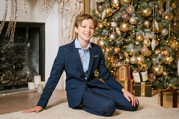 Caucasian boy in suit sits near christmas present. Cute teenager next to gift for christmas. Wait...