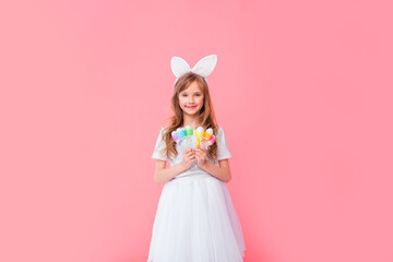 Obraz na płótnie Canvas Cute blonde girl 8-9 years old in a white T-shirt and skirt with rabbit ears holds Easter eggs isolated on a pink background. Happy Easter, holidays, traditions.