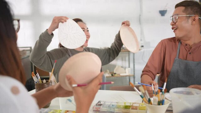 Group of Asian man and woman millennial generation friends enjoy and having fun painting self-made pottery together at pottery studio. Hobbies and leisure activity handmade ceramic painting workshop.