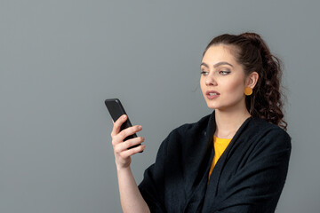 Fototapeta premium curly-haired emotional teenager woman in a black cloak using mobile phone, isolated on gray background