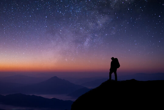 A young tourist with a bag is standing looking at the stars. The Milky Way and the beauty of the night sky alone on the top of the mountain. He felt successful at reaching his intended destination.