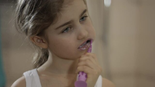 Close-up Portrait Young Little Girl Brushing Her Teeth in Bathroom Looking at Mirror Reflection. Daily Routine Dental Hygiene. Happy Portrait Cute Caucasian Toothless Child Brushing Teeth in Bathroom.