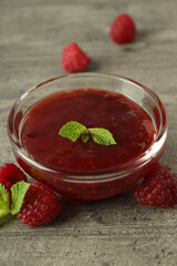 Glass bowl of raspberry jam with ingredients on gray textured background