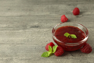 Glass bowl of raspberry jam with ingredients on gray textured background