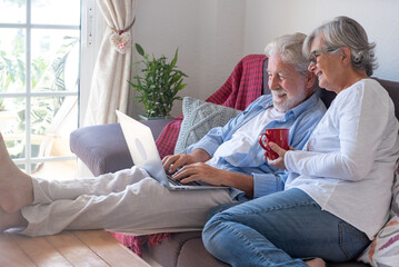 Relaxed senior couple at home sitting on sofa barefeet using laptop. Two elderly retirees enjoying...